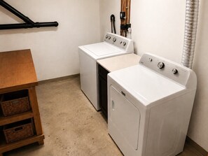 A laundry room with two white front-loading washers and dryers, a wooden table with baskets, and a black hose on the wall.