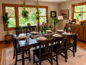 A dining area with a wooden table set for six, surrounded by chairs. There are potted plants, candles, and a decorative birdhouse on the table.