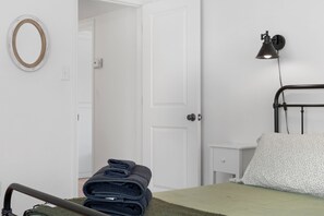 A neatly made bed with a green bedspread, a wall-mounted lamp, a bedside table, and a folded towel on the floor.