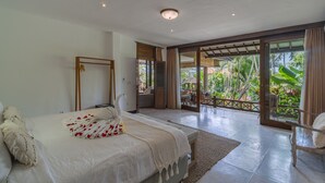 Quarto com cama, uma cadeira de madeira e vista para um jardim tropical através de uma porta de vidro.