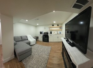 A modern living room with a grey sectional sofa, a flat-screen TV, a coffee machine, and a kitchen area with white cabinets and a wooden countertop. - Private 2BR Basement Suite / Patio•Cinema•Fire Pit (Sandy)