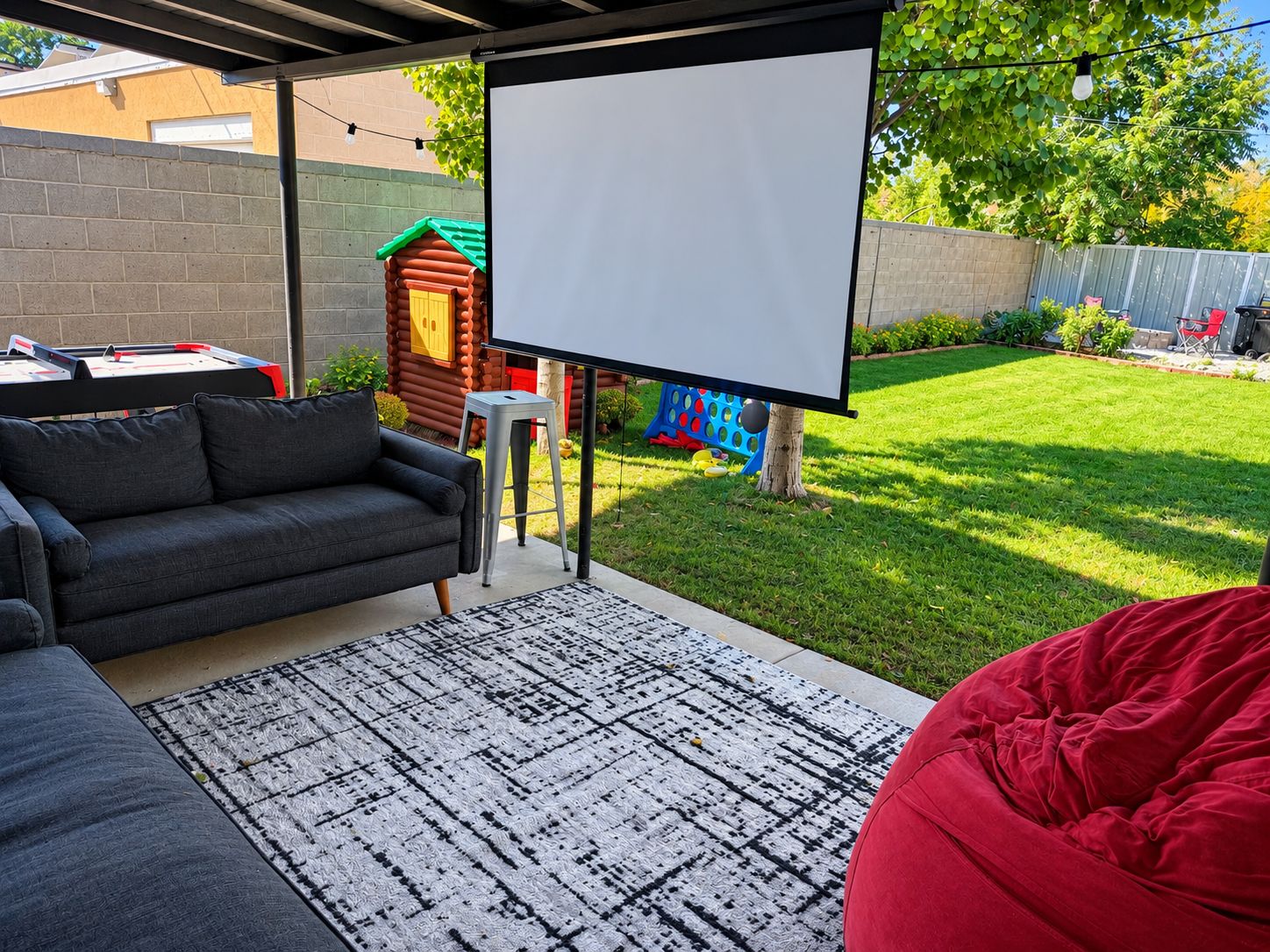 Eine Terrasse im Freien mit einer Couch, einem Sitzsack, einer Projektionsleinwand und einem kleinen Spielhaus im Hintergrund.