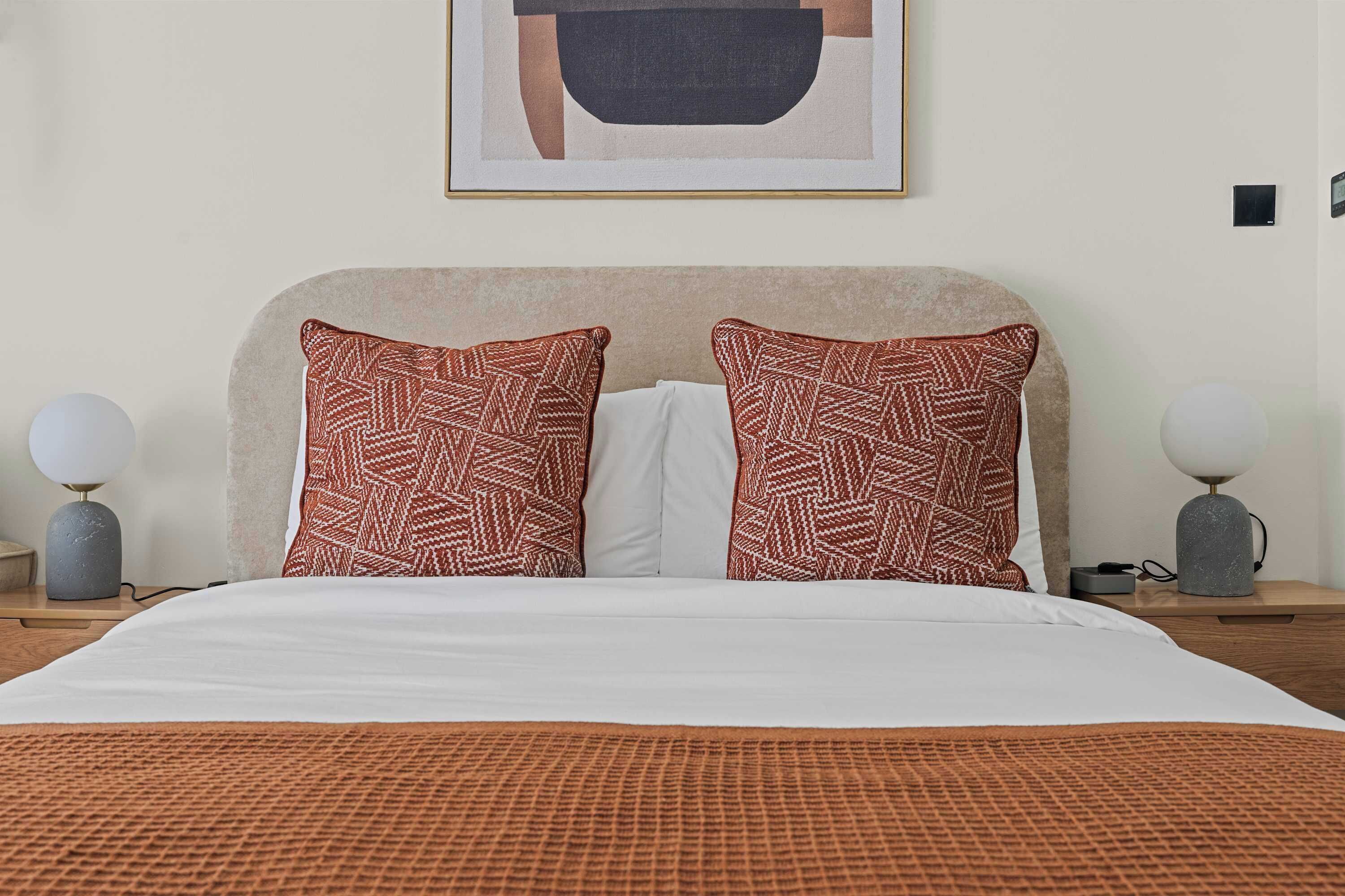 Una camera d'albergo con un letto, due cuscini decorati, un piumone bianco, un'opera d'arte incorniciata sopra il letto e due lampade ai comodini in legno.