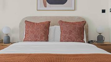 Una camera d'albergo con un letto, due cuscini decorati, un piumone bianco, un'opera d'arte incorniciata sopra il letto e due lampade ai comodini in legno.