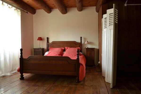 A wooden bed with a headboard, a nightstand with a lamp, and a wardrobe in a room with wooden flooring and a sloped ceiling.