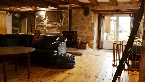 A rustic interior with exposed wooden beams, a stone wall, a fireplace, a leather sofa, and a wooden table.