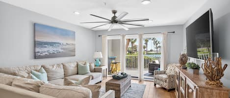 A modern living room with a ceiling fan, a large flat-screen TV, and a view of the outdoors through a sliding glass door.