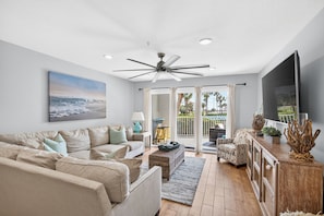 A modern living room with a ceiling fan, a large flat-screen TV, and a view of the outdoors through a sliding glass door. - Beautifully decorated 2 bed/2 bath w/ Beach Set up at Amalfi in Miramar Beach. (Miramar Beach)