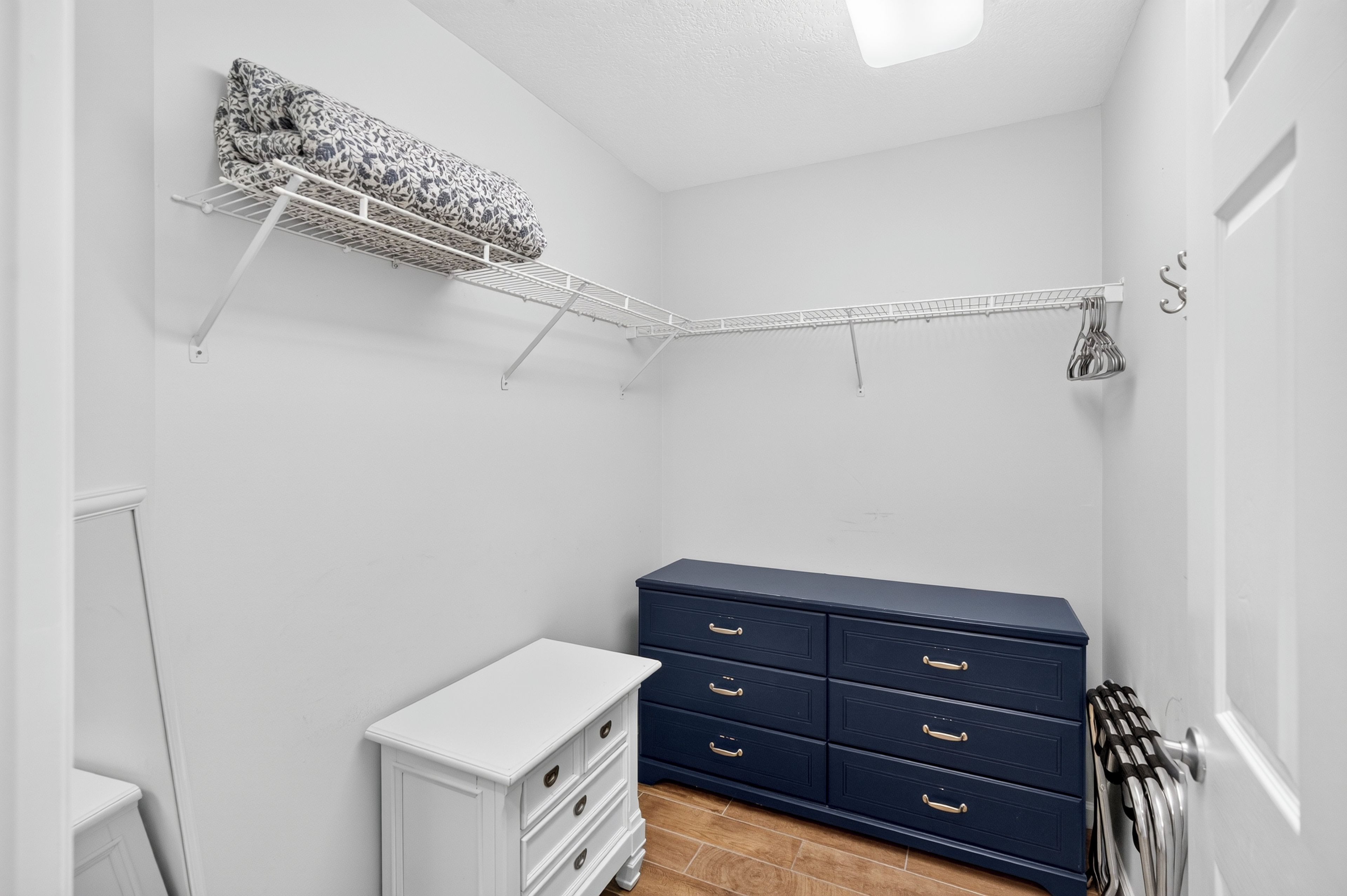 A small, well-lit room with a white dresser, a dark blue chest of drawers, a white cabinet, and a metal drying rack with clothes.