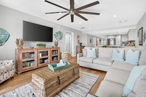 A modern living room with a ceiling fan, a flat-screen TV, a bookshelf, a sofa, and a kitchen in the background. - Beautifully decorated 2 bed/2 bath w/ Beach Set up at Amalfi in Miramar Beach. (Miramar Beach)