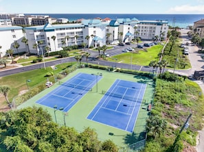 A tennis court with a green and blue surface, surrounded by a fence, with a view of a hotel and the ocean in the background. - Beautifully decorated 2 bed/2 bath w/ Beach Set up at Amalfi in Miramar Beach. (Miramar Beach)