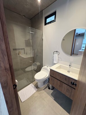 A modern bathroom with a glass-enclosed shower, a toilet, a sink with a mirror, and wooden cabinetry. - Balcony, birds, and nature · 2 bedrooms · Bogotá (Bogotá)