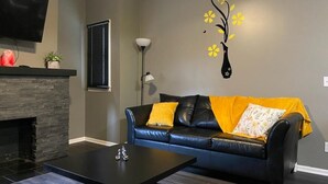 Ein Wohnzimmer mit Kamin, einem schwarzen Ledersofa mit gelben Kissen, einem flachbildfernseher über dem Kamin und einer Wandvase mit gelben Blumen.