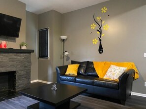 A living room with a fireplace, a black leather sofa with yellow cushions, a flat-screen TV mounted above the fireplace, and a wall-mounted vase with yellow flowers. - Walkable Little Italy Gem w/ King Bed Near CWRU (Cleveland)