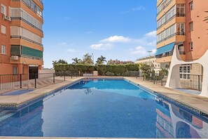 Une piscine à l’eau d’un bleu limpide, entourée d’une barrière basse et jouxtant des immeubles à plusieurs étages dotés de balcons.