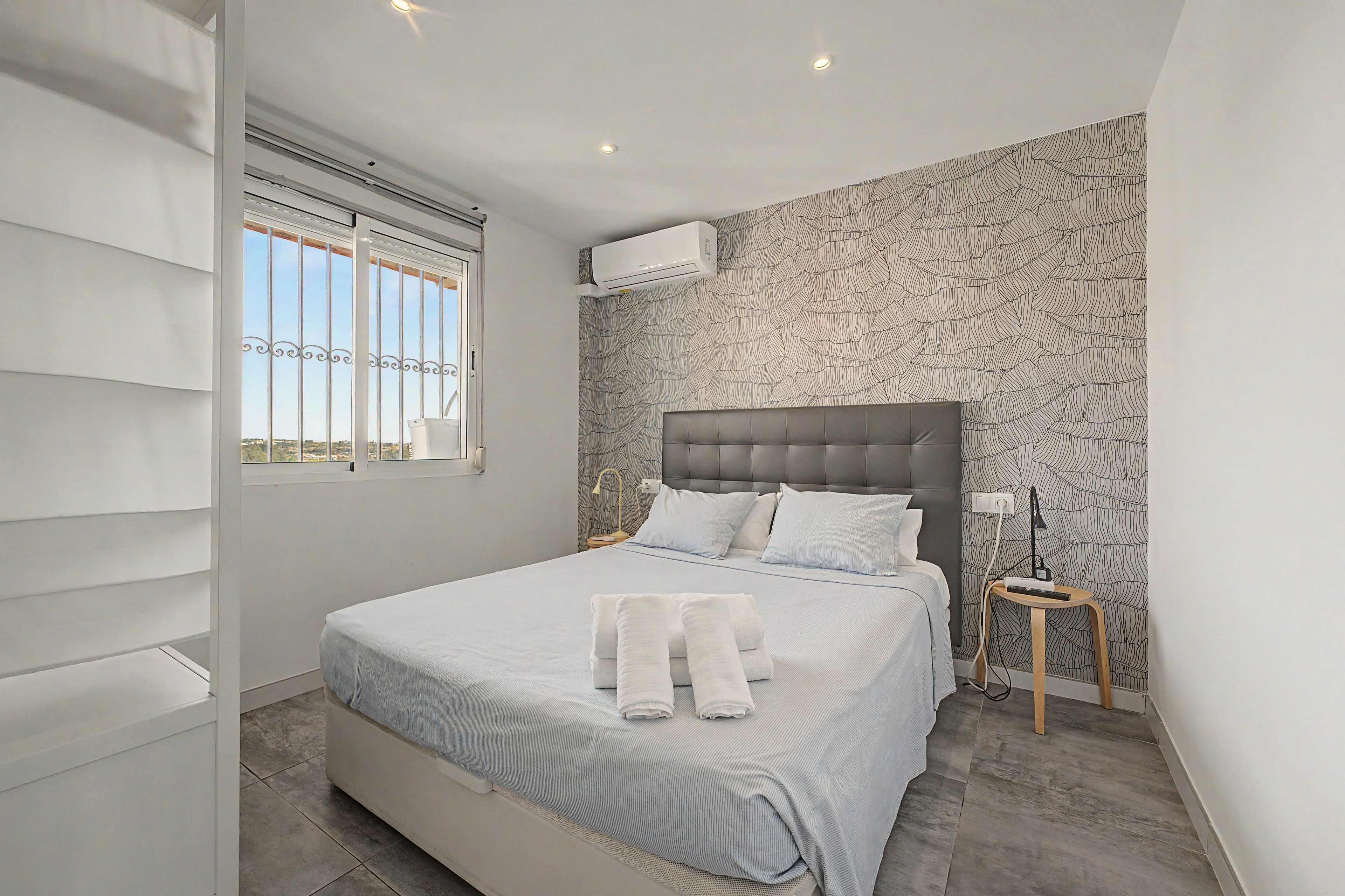 Une chambre à coucher avec un lit, une fenêtre, un climatiseur fixé au mur et un papier peint à motifs.