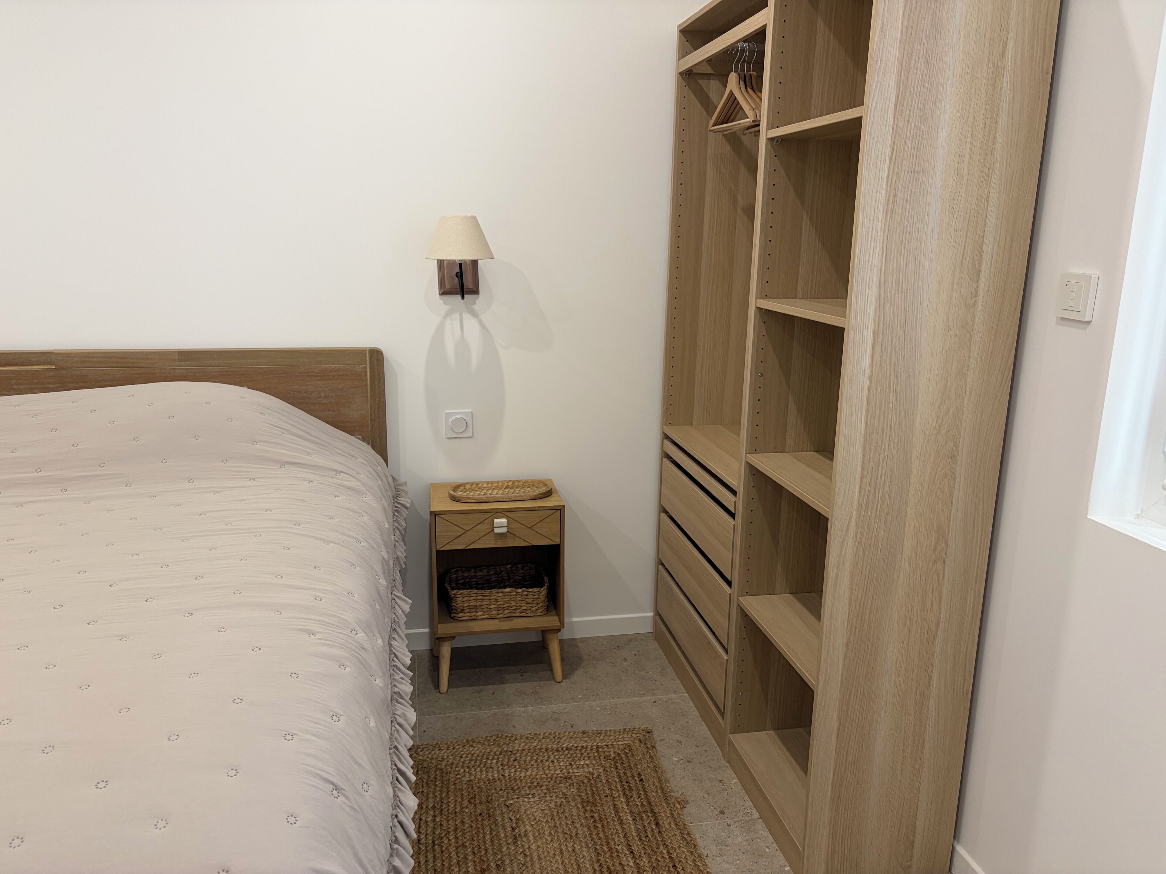 A bedroom with a bed, a nightstand, a lamp, a wooden wardrobe, and a woven rug.