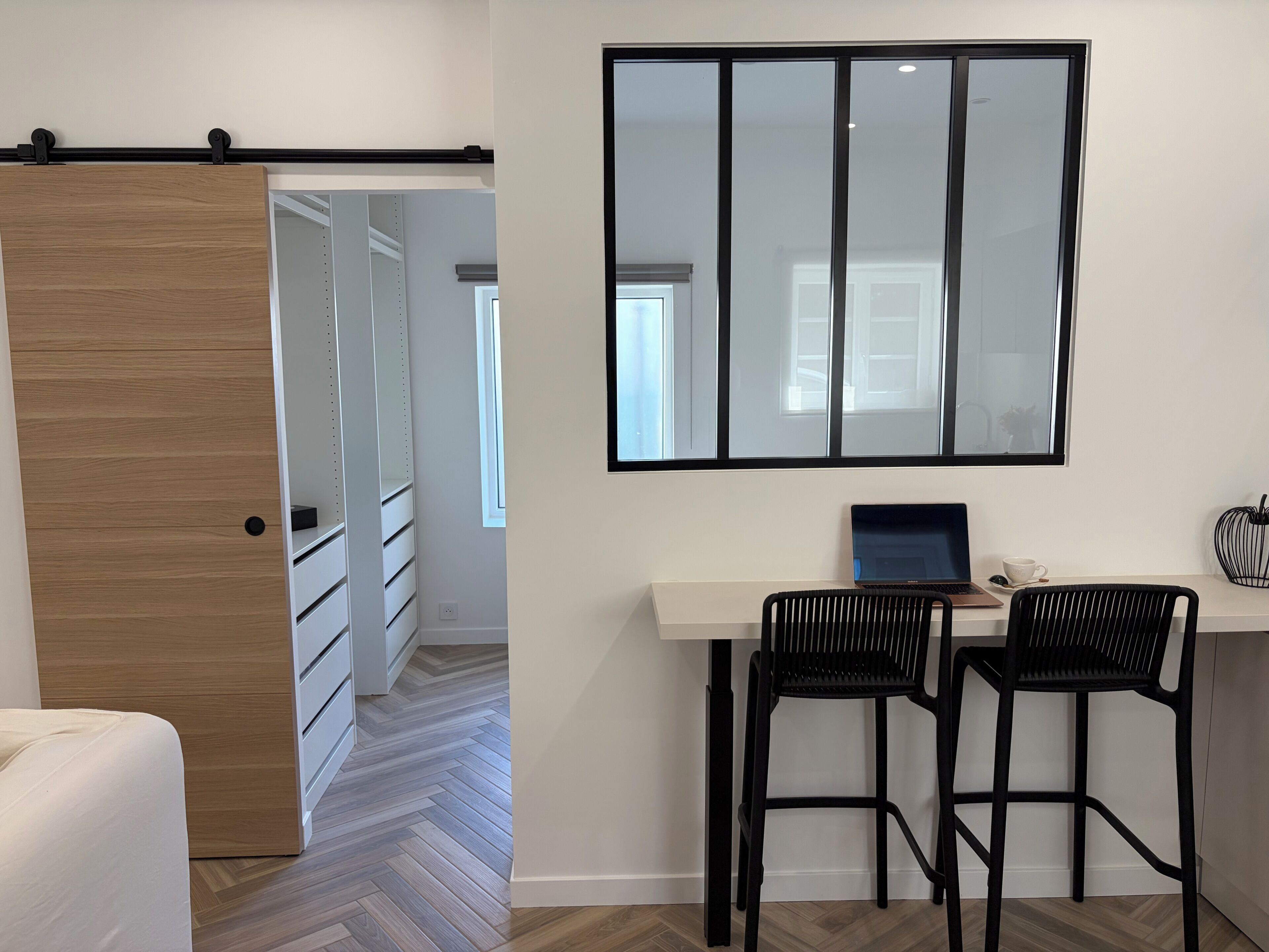 Une pièce moderne avec une porte en bois, un bureau blanc avec deux chaises noires, un ordinateur portable et un grand miroir.
