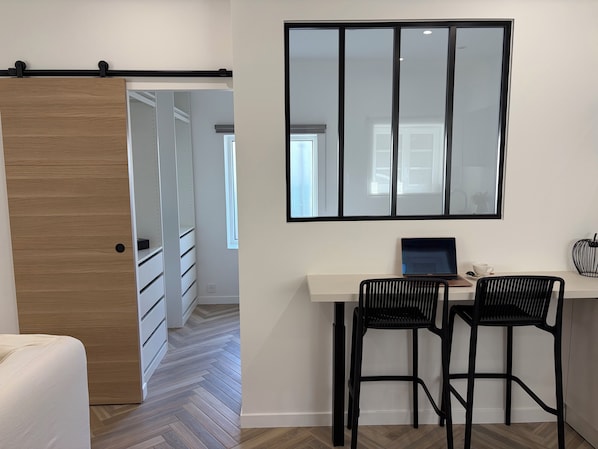 Una habitación moderna con una puerta de madera, un escritorio blanco con dos sillas negras, una computadora portátil y un espejo grande.