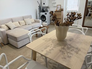 A modern living room with a sofa, a dining table, chairs, a washing machine, and a vase with dried flowers.