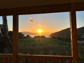 A sunset view from a wooden cabin with a deck, overlooking a grassy landscape and a body of water.