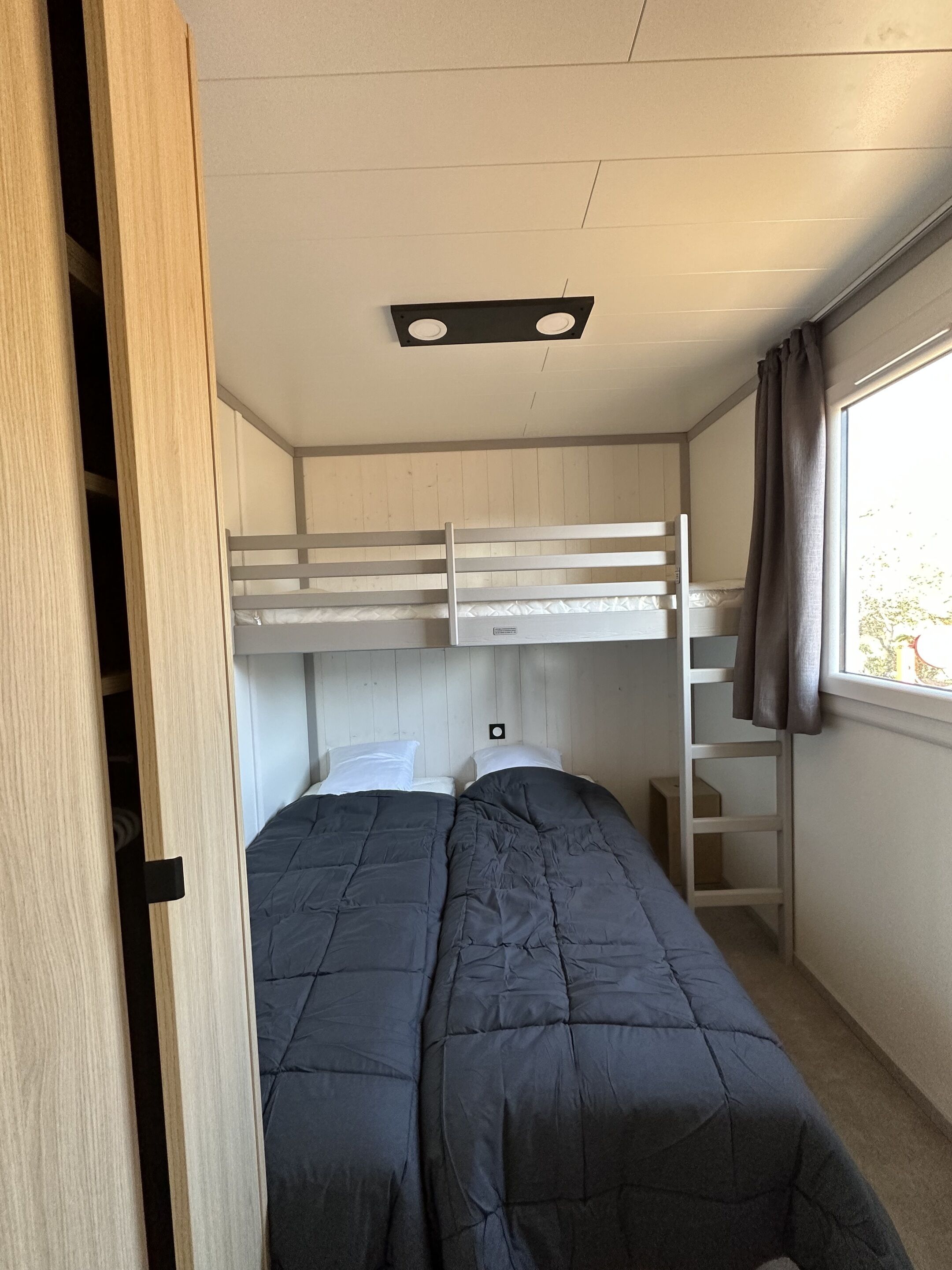 Een slaapkamer met stapelbed, een raam, een houten garderobe en een plafondlamp.