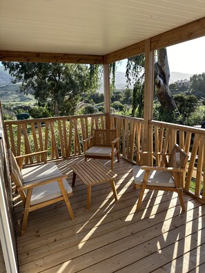 A wooden deck with a covered area, furnished with two chairs and a small table, overlooking a landscape with trees and a distant house.