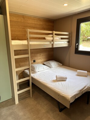 A bunk bed with a wooden headboard, a single bed below, white bedding, and a window with a view of greenery.
