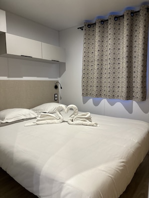 A hotel room with a neatly made bed, a headboard, a wall-mounted light, and a patterned curtain.