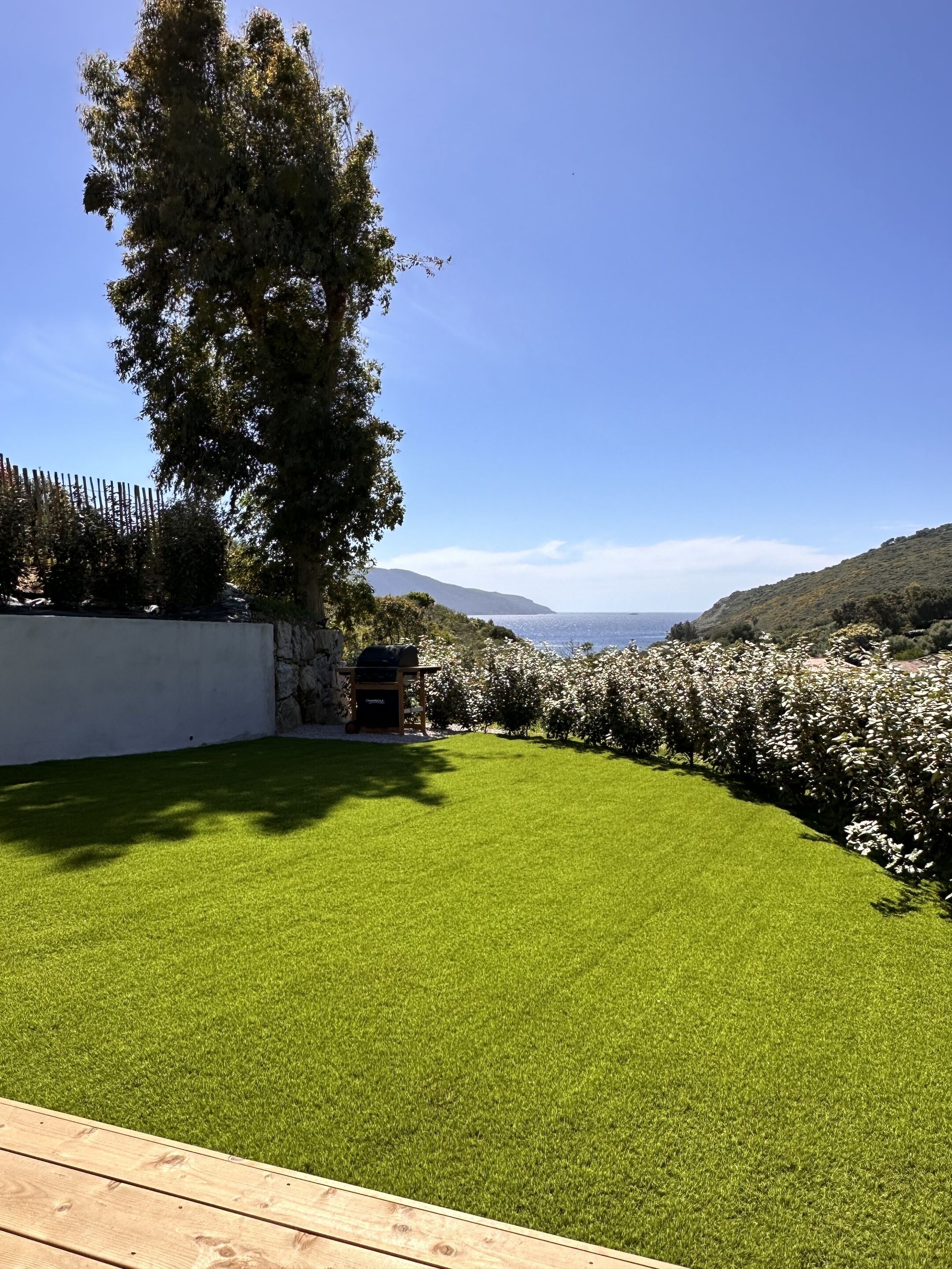 A well-maintained lawn with a wooden deck, a barbecue grill, and a view of the sea and hills.