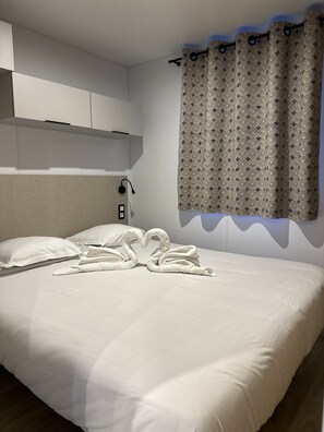 A hotel room with a neatly made bed, a headboard, a wall-mounted light, and a patterned curtain. - Les Sanguinaires - Mobile Home (Appietto)