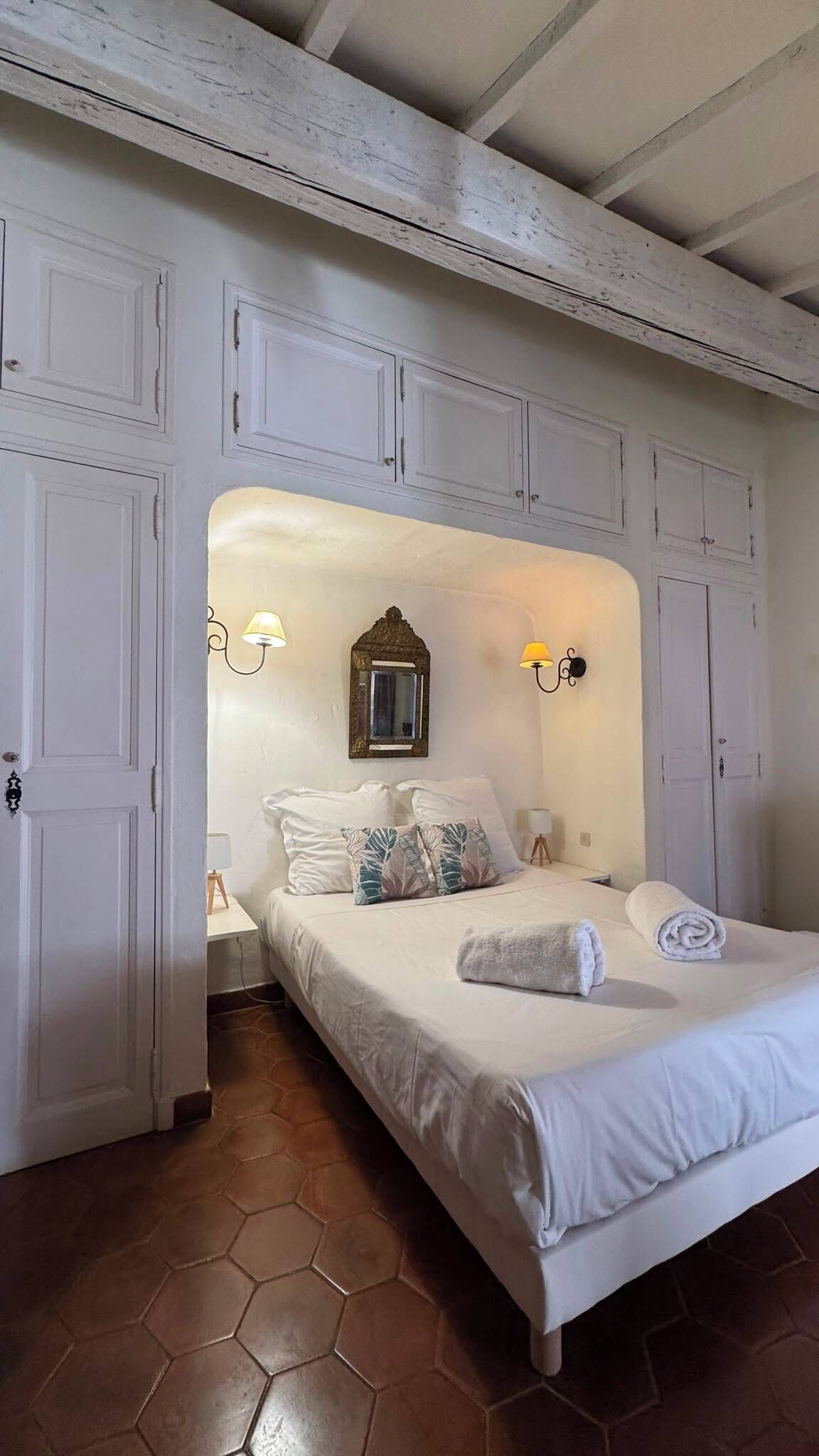 Een slaapkamer met een bed, witte wanden, een vloer van honingraat tegels en een plafond met zichtbare balken.