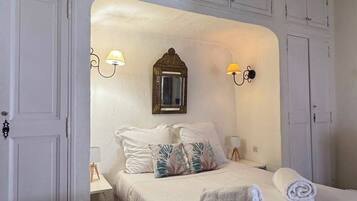 Een slaapkamer met een bed, witte wanden, een vloer van honingraat tegels en een plafond met zichtbare balken.