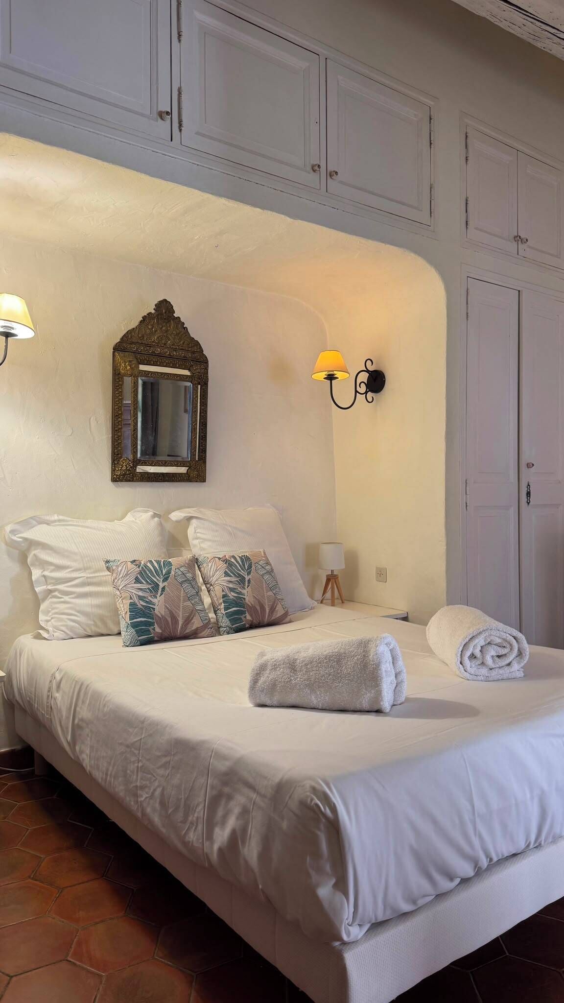 A neatly made bed with white linens and a rolled towel, a wall-mounted mirror with an ornate frame, and two wall sconces with lamps.