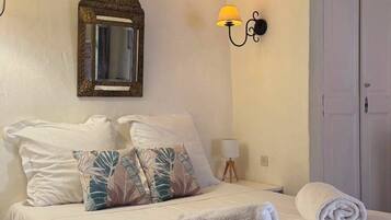 A neatly made bed with white linens and a rolled towel, a wall-mounted mirror with an ornate frame, and two wall sconces with lamps.