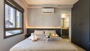 Habitación de hotel moderna con una cama grande, una mesita de noche con lámpara, una ventana con vistas a edificios y un equipo de aire acondicionado en la pared.