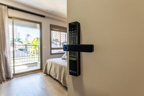 A modern room with a sliding glass door leading to a balcony, a bed with a white bedspread, and a black wall-mounted control panel. - GA 45 Ape com AR prox. Allianz  (São Paulo)