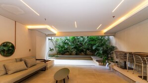 Un interior moderno con un sofá, una mesita redonda y una escalera que conduce a un balcón con abundante vegetación.