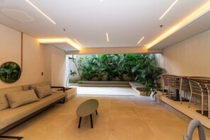 Um interior moderno com um sofá, uma mesa redonda pequena e uma escadaria que leva a uma varanda com vegetação exuberante.