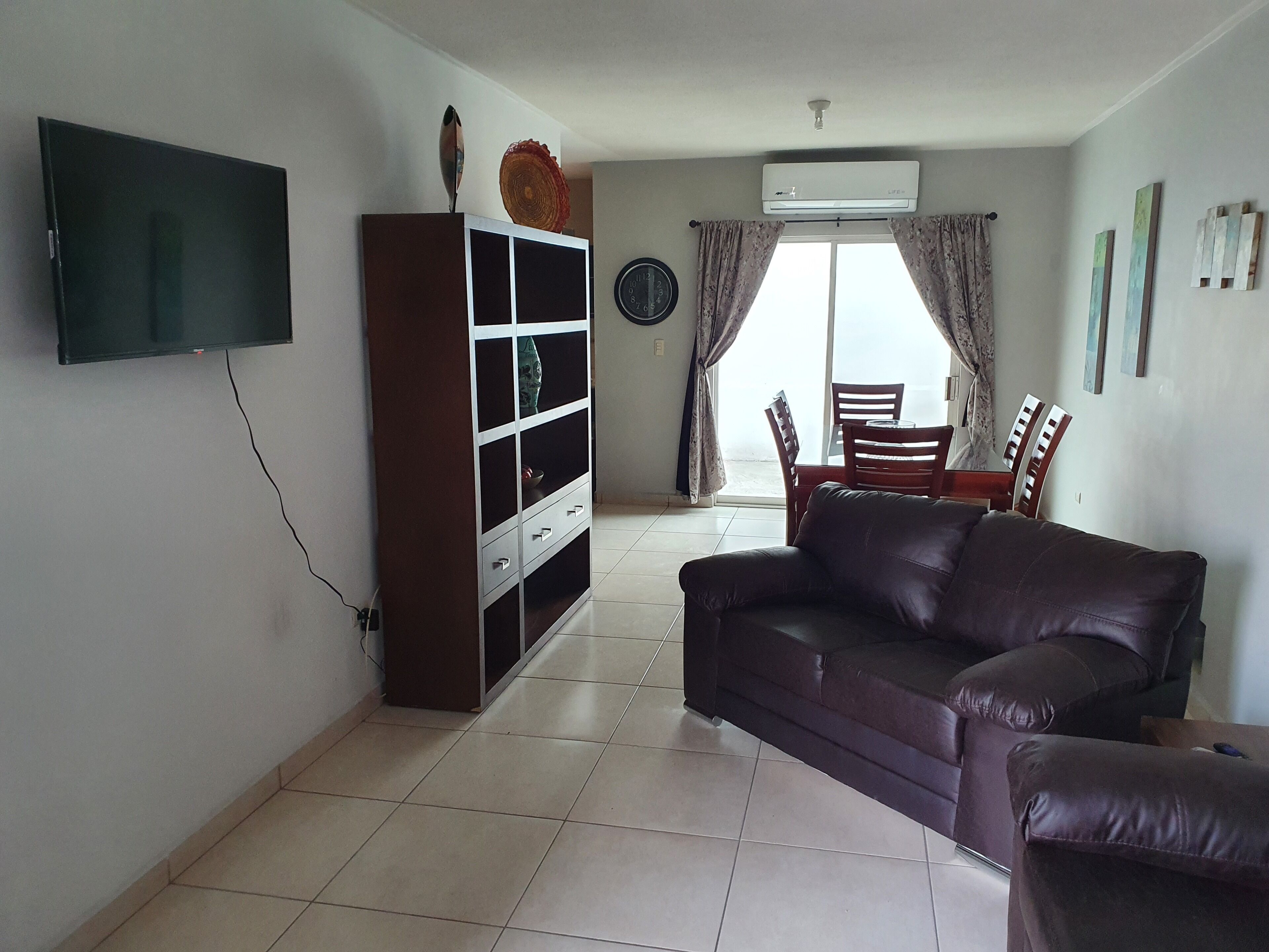 A living room with a flat-screen TV, a brown sofa, a wooden cabinet, a dining area with chairs, and a large window with curtains.