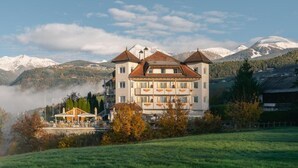 A large building with multiple floors and a central tower, surrounded by a green landscape and snow-capped mountains in the background.