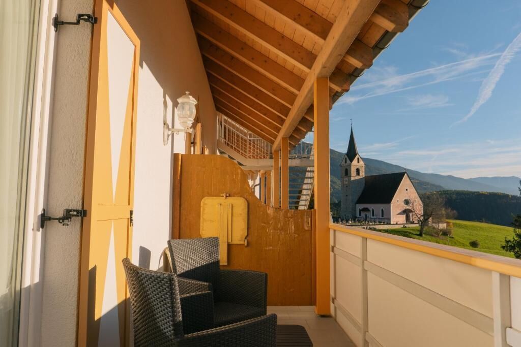 Un balcón con mobiliario de mimbre, techo de madera y vistas a una iglesia y montañas.