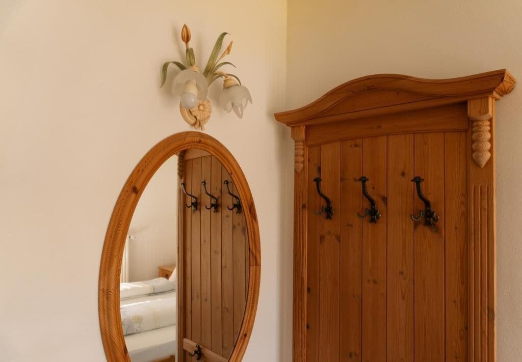 Un armario de madera con cuatro ganchos negros, un espejo con marco de madera y una lámpara de pared con dos flores.