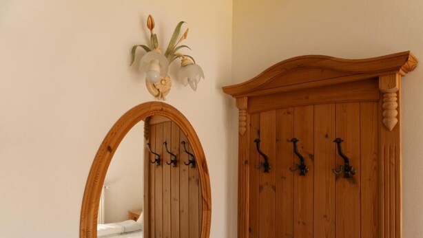 A wooden wardrobe with four black hooks, a mirror with a wooden frame, and a wall-mounted light fixture with two flowers.