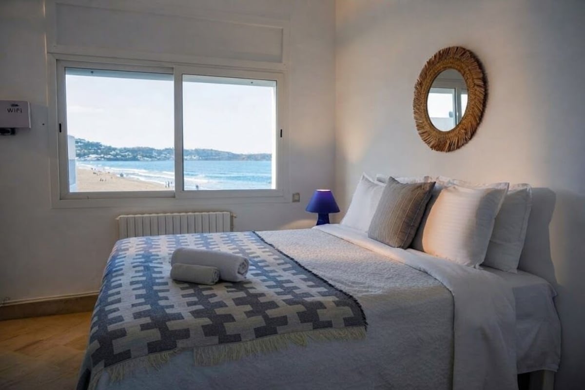 Een slaapkamer met een bed, een raam met uitzicht op het strand, een spiegel aan de muur en een blauwe lamp.