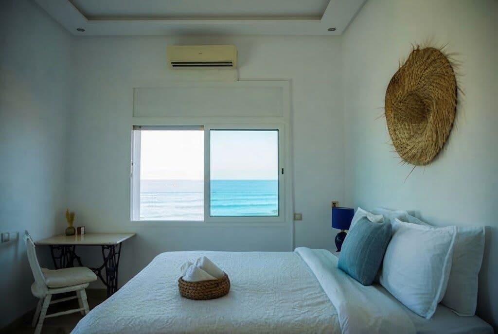 A bedroom with a bed, a chair, a small table, a window with a view of the sea, and a woven wall decoration.