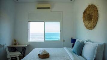 A bedroom with a bed, a chair, a small table, a window with a view of the sea, and a woven wall decoration.