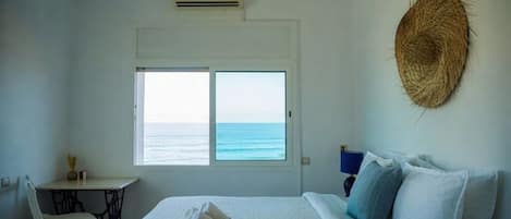 Un dormitorio con cama, una silla, una mesita, un ventanal con vista al mar y un adorno de pared tejido.