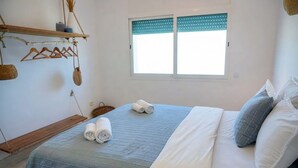 Un dormitorio con cama, almohadas, ventana con persianas, estante con objetos colgando y un perchero.