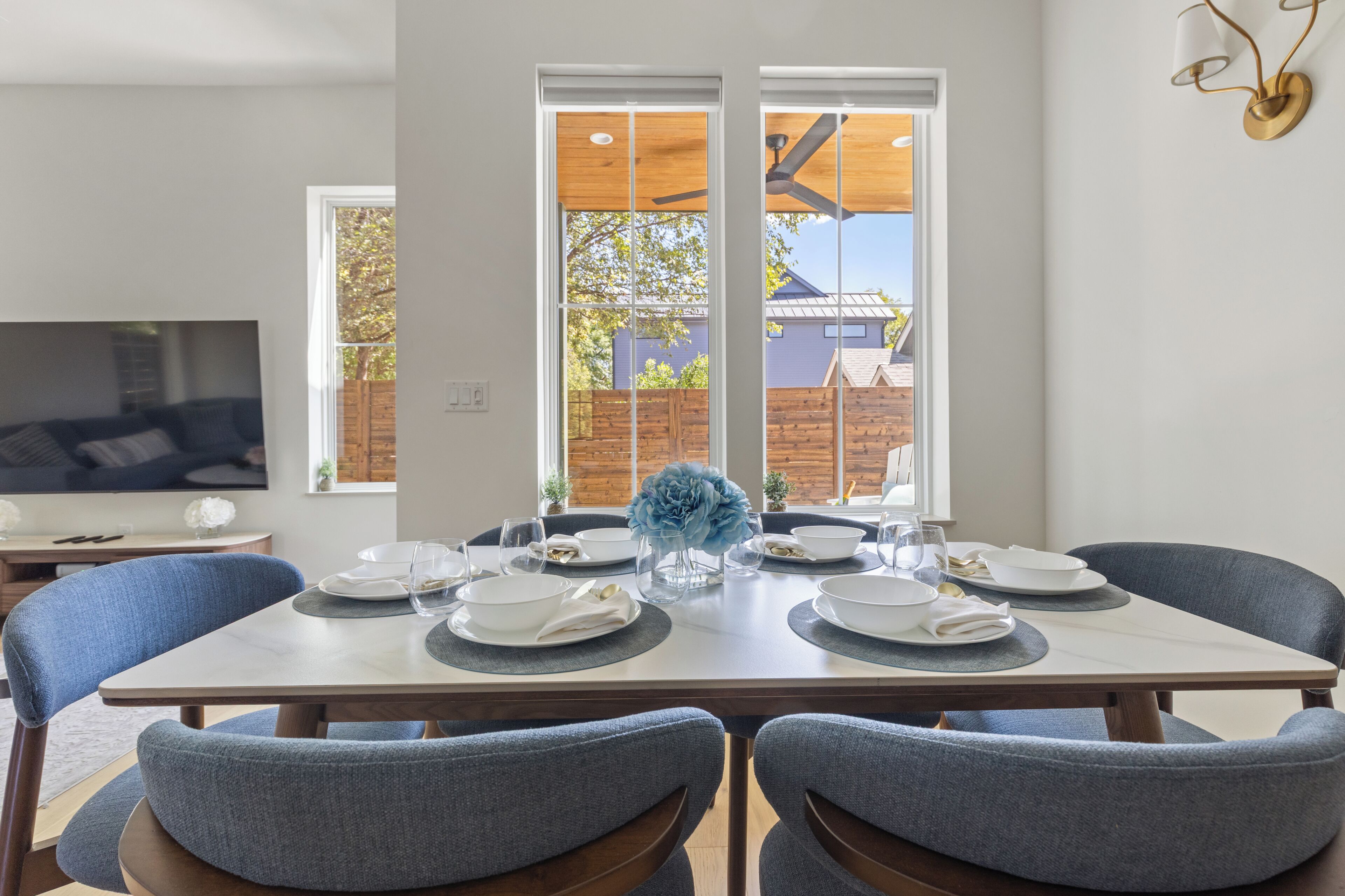 A dining area with a table set for four, chairs, a television, and a view of a backyard through large windows.